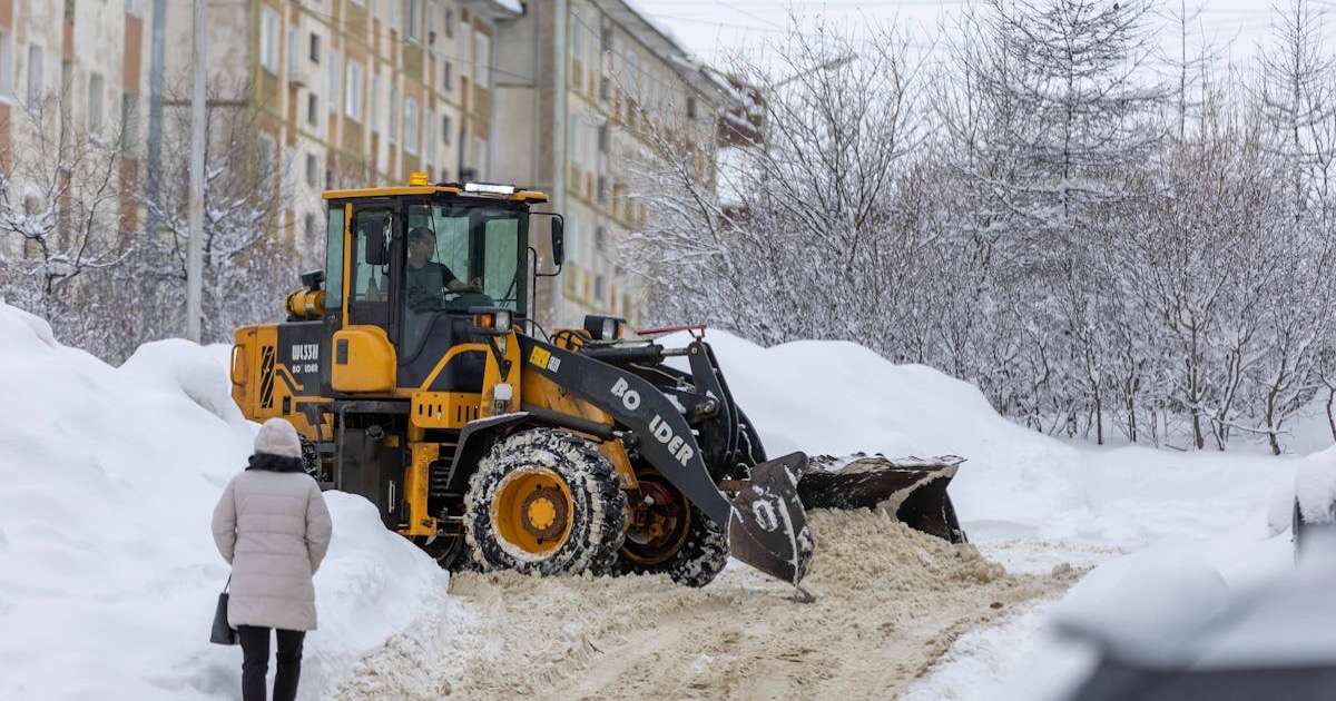 Yellow tractor clearing snow on a winter street near residential buildings.