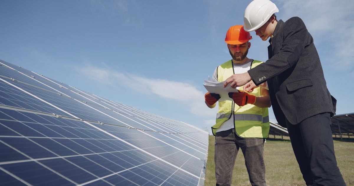Low angle of young inspector and foreman in hardhats checking documentation against modern solar panels in field