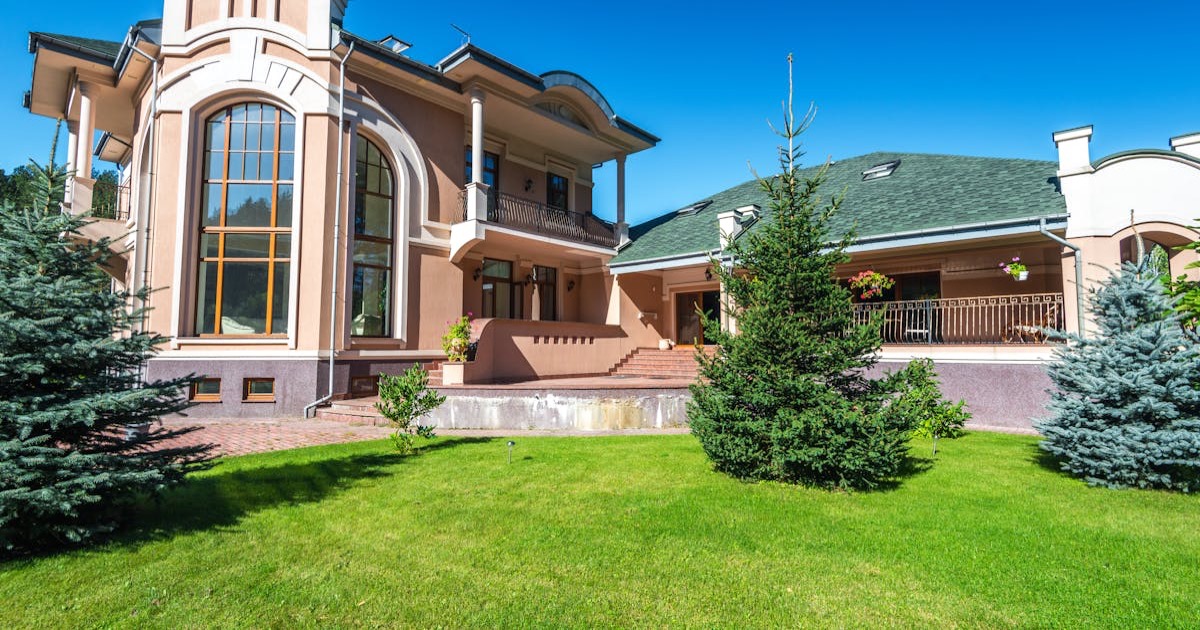 Modern luxury villa with large windows, surrounded by lush green lawn and trees.